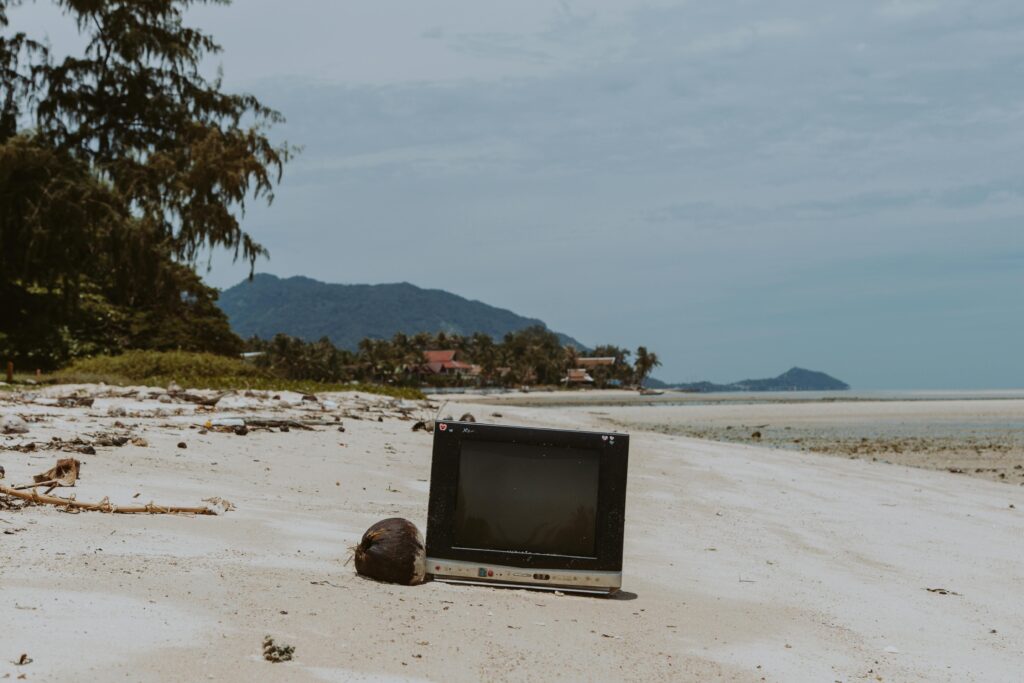 An old television placed on a deserted sandy beach with coconut and palm trees.