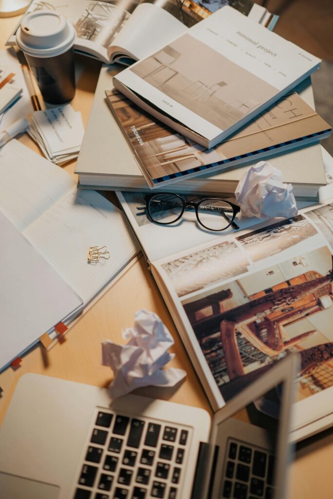 A cluttered desk with books, eyeglasses, crumpled papers, and a laptop, creating an unorganized workspace.