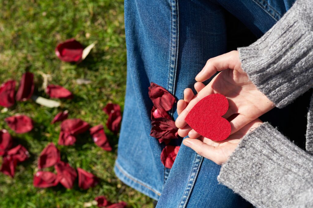 A woman sits on grass holding a red heart-shaped card, surrounded by rose petals, symbolizing love and compassion.