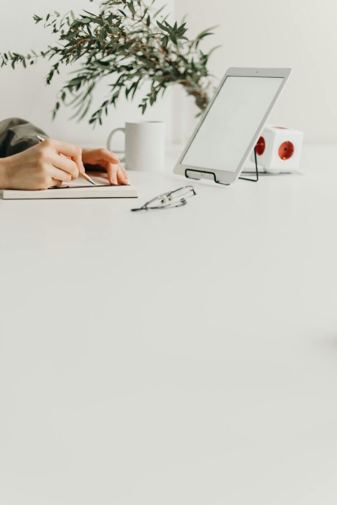 A clean, minimalist workspace with a tablet, plant, and hand writing in a notebook.