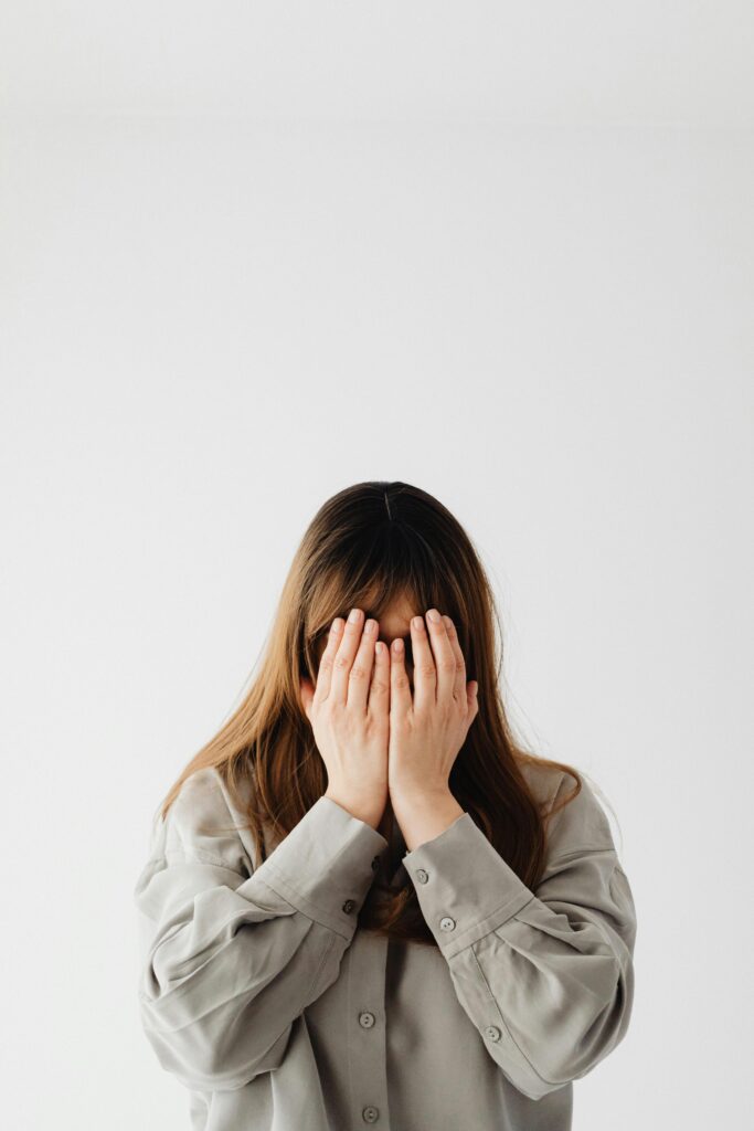 A shy young woman with long brunette hair covers her face in a light-colored room, conveying emotion indoors.